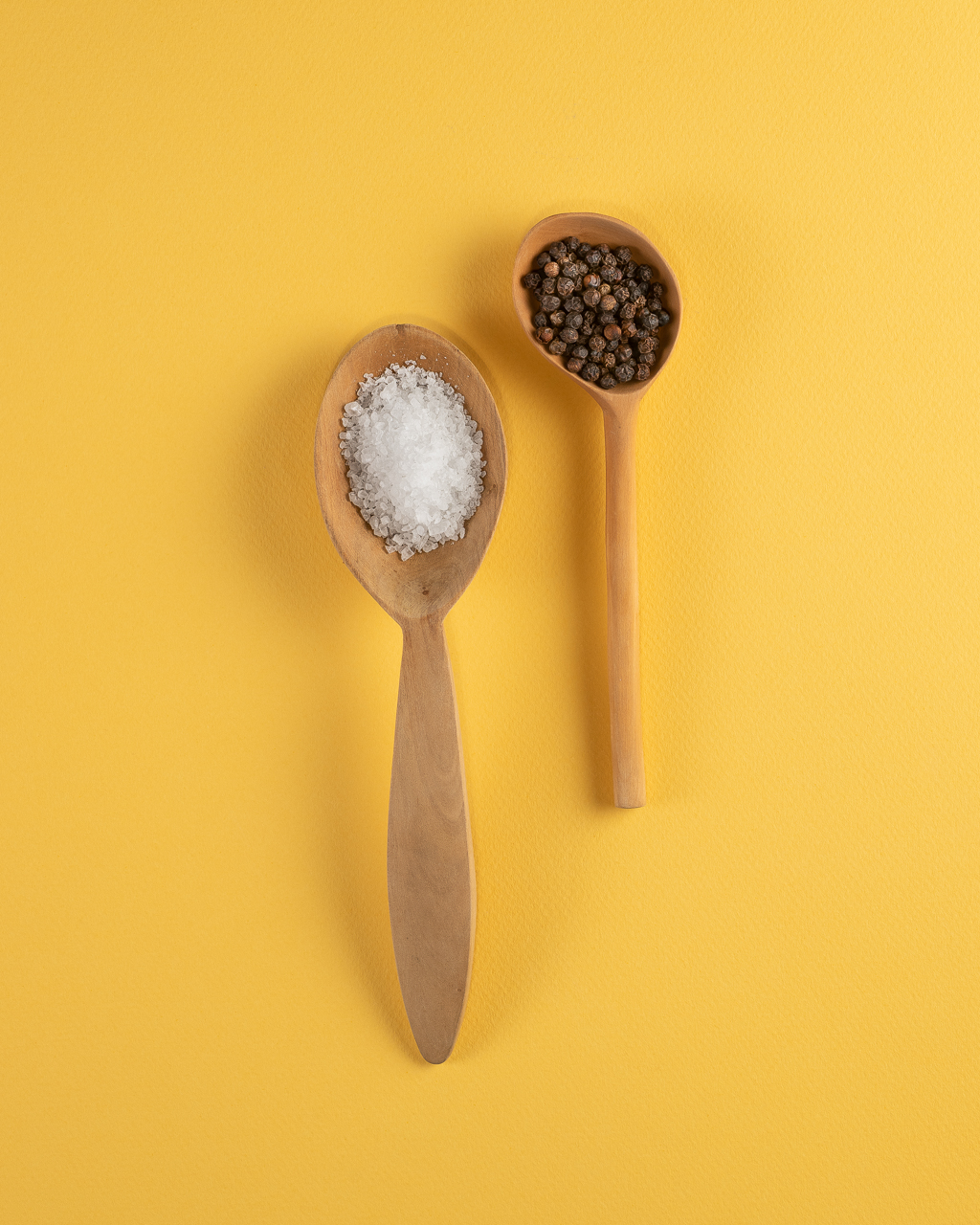 Salt and pepper on wooden spoons on yellow by Jose Barrios